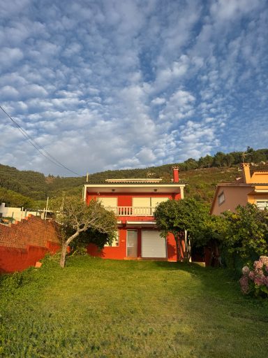 Fachada principal Casa roja con jardín verde, al fondo montañas y cielo nublado.